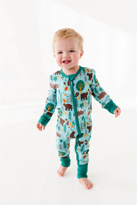 Baby model stands wearing blue green bamboo pajama in Woody Woodland Bear print with mountains and mushrooms