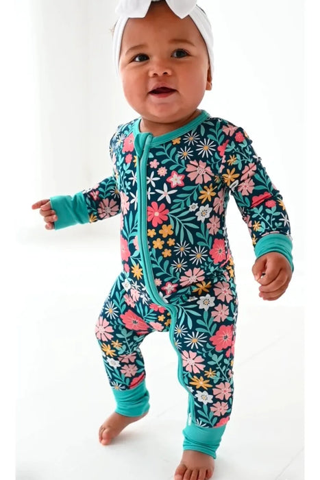 Smiling baby model standing in Maren Floral One Piece Zip Sleep Romper with pink flowers on navy background.