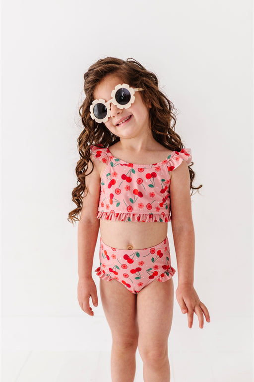 Child wearing a red cherry-themed two-piece swimsuit with sunglasses on a white background Bird and Bean Kids 