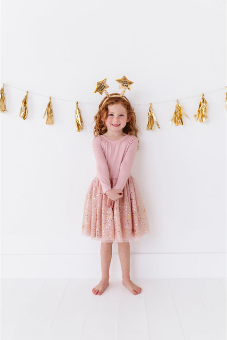 Young girl in a pink sparkly dress standing against a white wall with gold tassel decorations. | Bird and Bean 