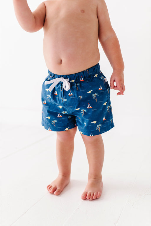 Child wearing blue swim shorts with a palm tree and sailboat pattern on a white background Bird and Bean Kids