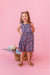Young girl in a floral bamboo fabric dress standing next to a basket of flowers on a pink background | Bird and Bean Kids