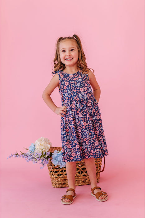 Young girl in a blue and pink  floral bamboo fabric tank dress standing against a pink background with flowers and a basket. | Bird and Bean Kids