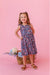 Young girl in a blue and pink  floral bamboo fabric tank dress standing against a pink background with flowers and a basket. | Bird and Bean Kids