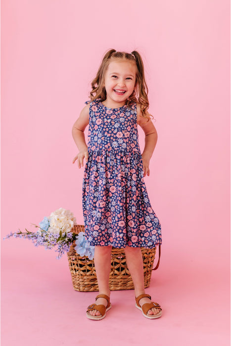 Young girl in a floral  bamboo fabric dress standing against a pink background with flowers and a basket. Bird and Bean Kids 