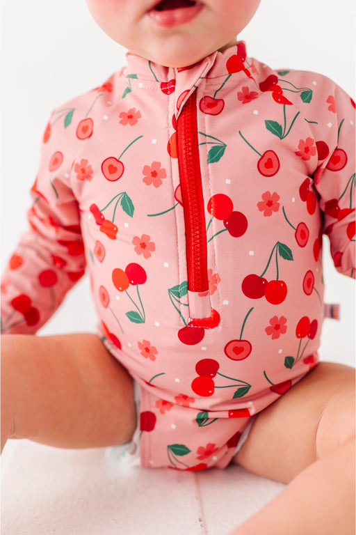 Baby wearing a pink rashguard zip front swimsuit with cherry pattern - Bird and Bean Kids