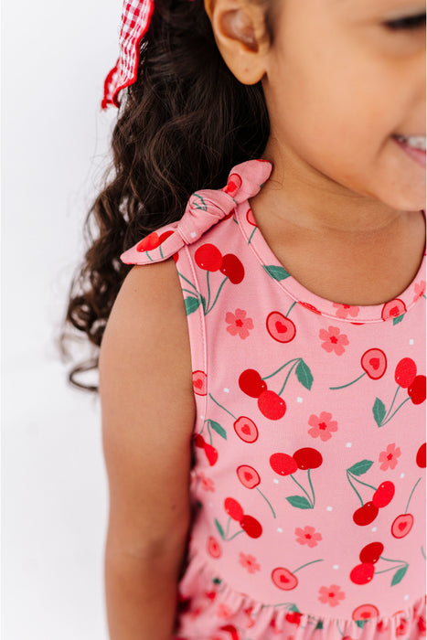 Child wearing a pink dress with bow shoulder tie with cherry pattern on a white background | Bird and Bean Kids