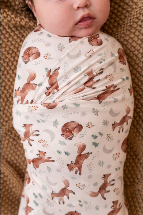 A close-up of a baby wrapped in a soft bamboo swaddle blanket featuring a woodland print of brown foxes, crescent moons, and small leaves on a cream background, resting on a textured brown blanket. Bird and Bean Baby 