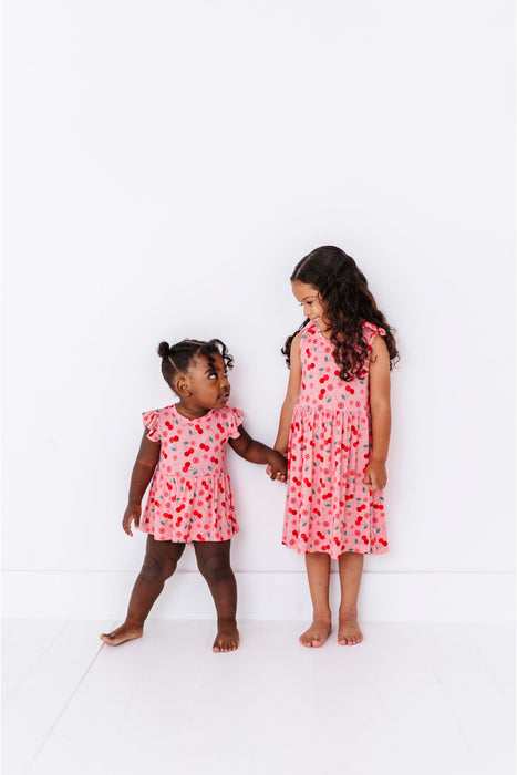 Two young girls wearing matching pink cherry print bamboo fabric dresses with a floral pattern on a white background - Bird and Bean Kids