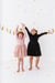 Two children in dresses standing with arms raised against a white background with gold confetti. | Bird and Bean Holiday Shop