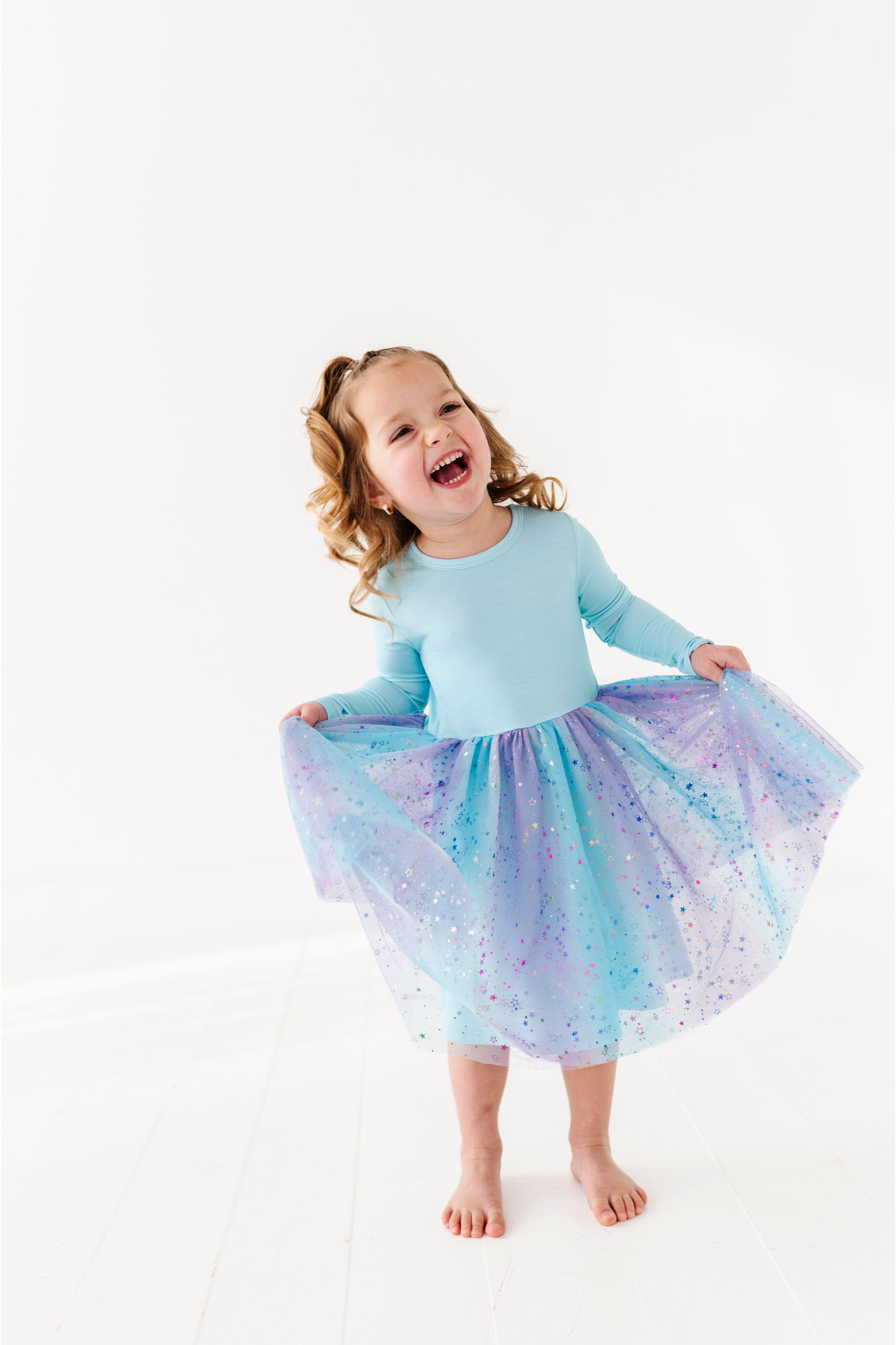 A young girl with curly light brown hair stands barefoot on a white studio floor, smiling and twirling with a light blue bamboo  long-sleeve top and a sparkly tulle tutu skirt in pastel blue, lavender, and purple. Bird and Bean Kids
