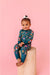 Baby sitting on a wooden block against a pink background wearing a floral zip pajamas | Bird and Bean 