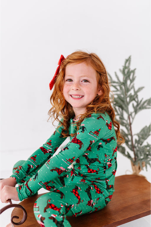 Young girl wearing green pajamas with red dog holiday pattern, sitting on a wooden stool. Bird and Bean original