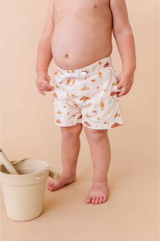 Child wearing patterned  swim shorts standing next to a white bucket on a beige background Bird and Bean