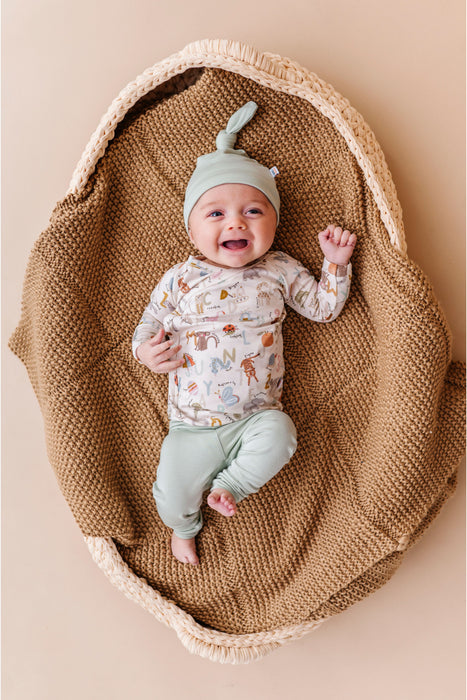 Baby in a woven basket wearing a patterned onepiece and green hat on a beige background. Bamboo Take Me Home Set – Animal ABC Bird and Bean Original