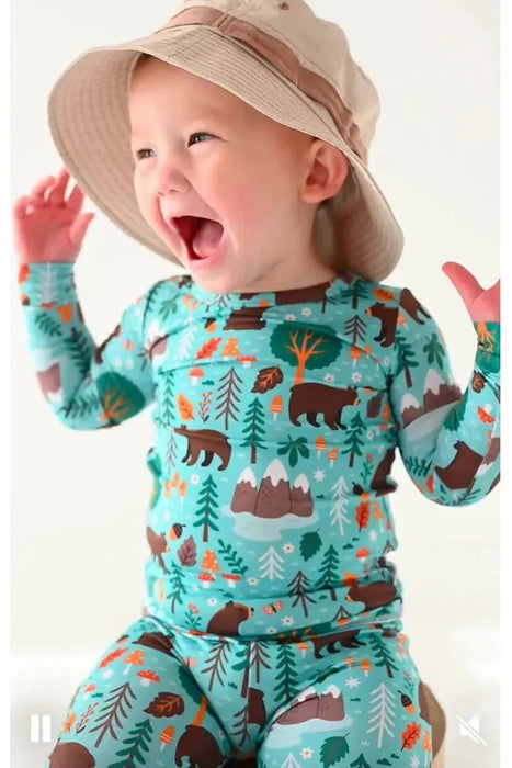 Boy model laughs while wearing blue green bamboo pajama set in Woody Woodland Bear print with mountains and mushrooms