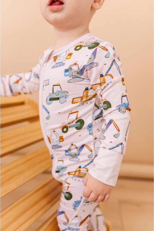 Child wearing a white long-sleeve shirt with colorful construction vehicle pattern on a wooden floor. Bamboo PJs by Bird and Bean Kids