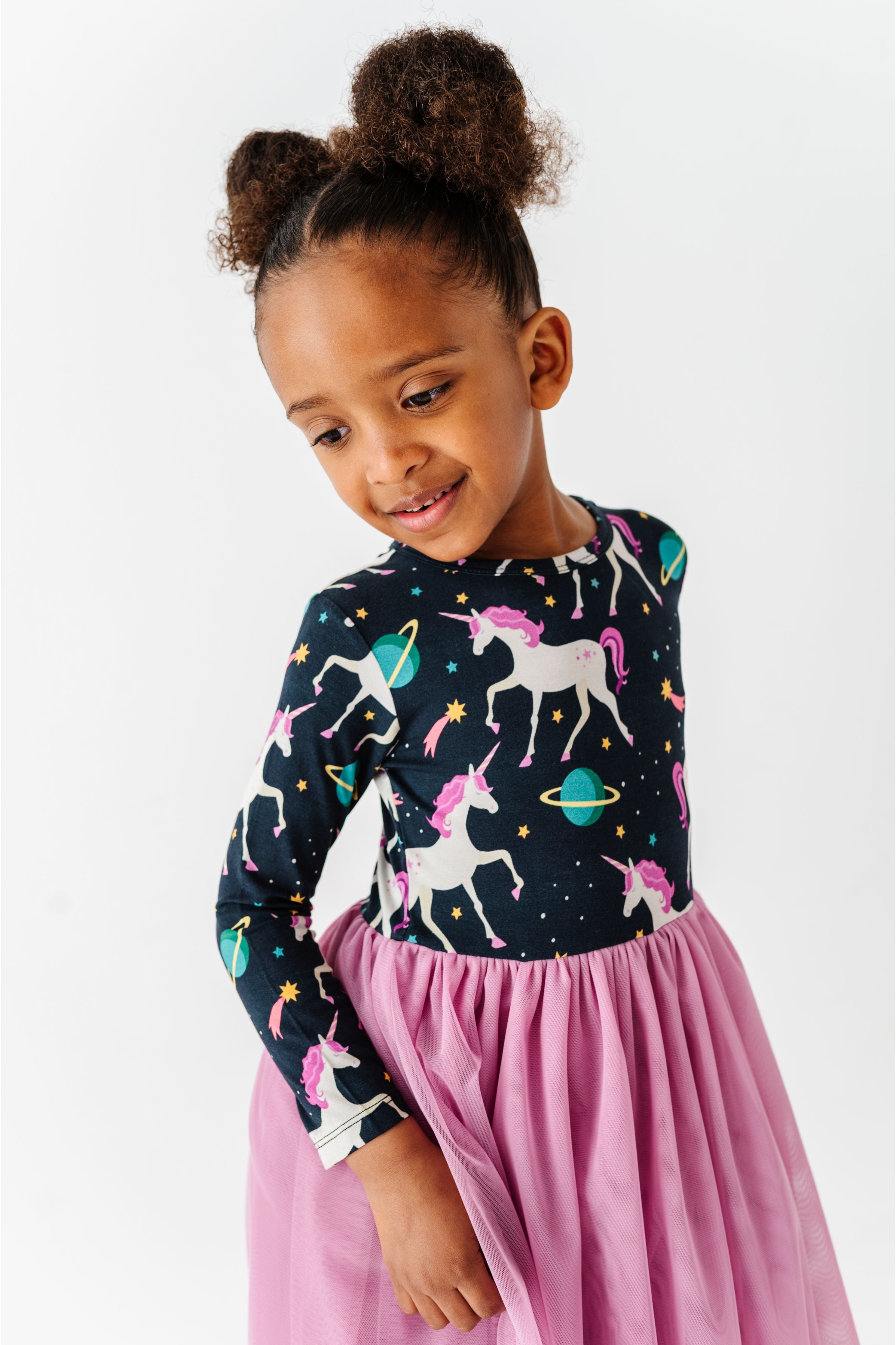 A young girl with her hair in two buns is wearing a long-sleeve top and a pink skirt. The top has a dark navy blue background with a colorful print of white unicorns, pink and teal starry accents, and small planets. Bird and Bean Tulle Dresses 