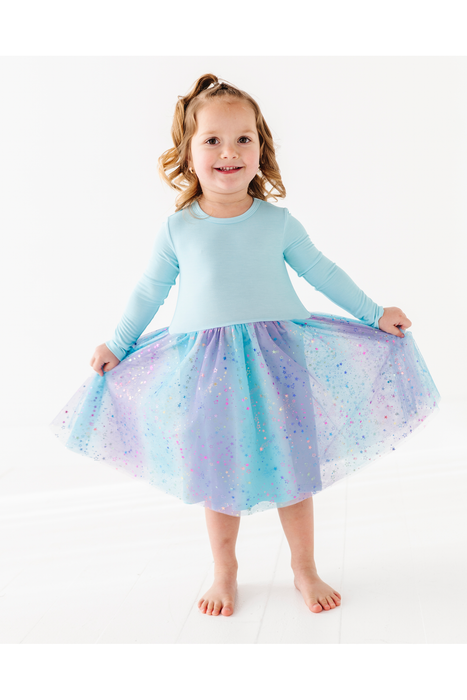 A young girl with blonde hair in a ponytail stands barefoot on a white studio floor, wearing a light blue long-sleeve top and a sparkly tulle tutu skirt in pastel blue, purple, and pink. She is holding out the sides of the skirt to show its volume. Bird and Bean Kids