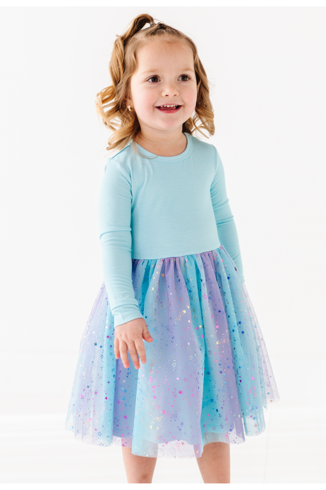 A young girl with blonde hair in a ponytail is wearing a light blue long-sleeve top and a light blue, purple, and pink glittery tulle skirt, standing against a plain white background. Bird and Bean Kids