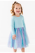 A young girl with blonde hair in a ponytail is wearing a light blue long-sleeve top and a light blue, purple, and pink glittery tulle skirt, standing against a plain white background. Bird and Bean Kids