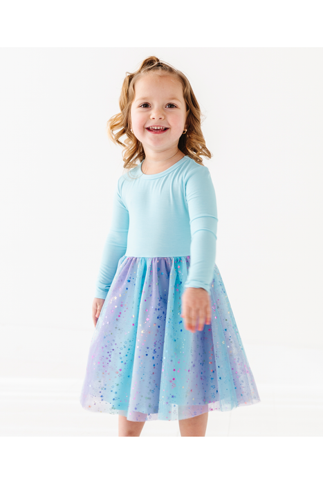 A young girl with blonde hair wearing a light blue long-sleeve top and a sparkly blue, purple, and pink tulle skirt, standing against a plain white background. Bird and Bean Exclusive Dress