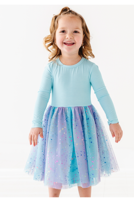 A young girl with blonde hair in a ponytail is smiling and wearing a light blue long-sleeve top with a sparkly tulle skirt in shades of blue, purple, and pink, set against a plain white studio background. Bird and Bean Girls Tulle Dresses