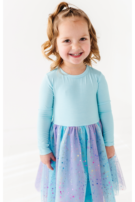 A young girl with blonde hair in a ponytail smiles while wearing a light blue long-sleeve top and a sparkly blue, purple, and pink tulle skirt against a white background. Bird and Bean exclusive design. 