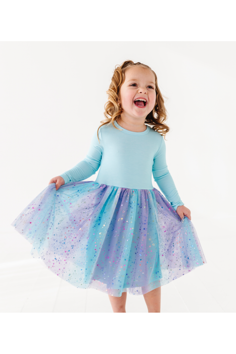 A young girl with curly blonde hair is smiling and laughing while twirling, wearing a light blue long-sleeve top and a voluminous tulle tutu skirt with a blue, purple, and pink gradient and glittery speckles. She is posing on a clean white studio background. Bird and Bean Dresses