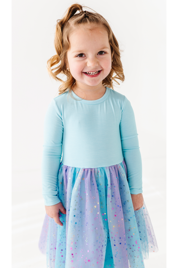 A young girl with blonde hair in a ponytail smiles while wearing a light blue long-sleeve top and a sparkly blue, purple, and pink bamboo tulle skirt against a white background. Bird and Bean Kids