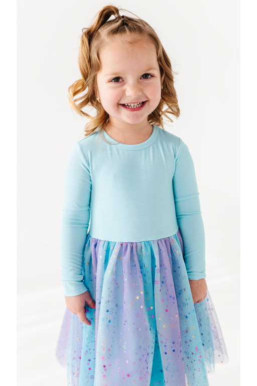 A young girl with blonde hair in a ponytail smiles while wearing a light blue long-sleeve top and a sparkly blue, purple, and pink bamboo tulle skirt against a white background. Bird and Bean Kids