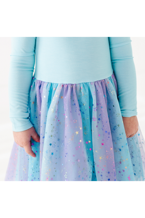 A close-up of a child wearing a light blue long-sleeve top and a pleated tulle skirt in pastel blue, lavender, and purple with scattered metallic star confetti. Bird and Bean Tulle Dresses