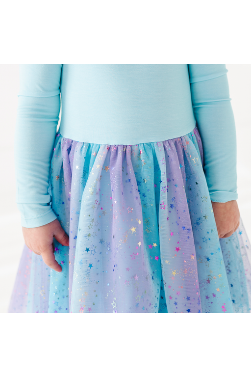 A close-up of a child wearing a light blue long-sleeve top and a pleated tulle skirt in pastel blue, lavender, and purple with scattered metallic star confetti. Bird and Bean Tulle Dresses