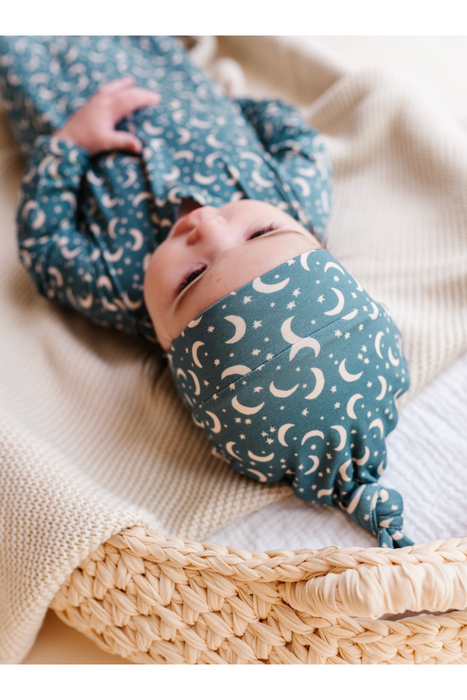 A baby lying on a cream knit blanket in a woven basket, wearing a matching long-sleeve outfit and knotted head covering with a bamboo blue-gray and cream crescent moon and star pattern. Bird and Bean Baby 