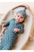 A baby lying on a textured cream blanket inside a woven basket, wearing a matching blue and white crescent moon patterned footless  bamboo sleeper and knotted beanie. Bird and Bean Baby Gifts 