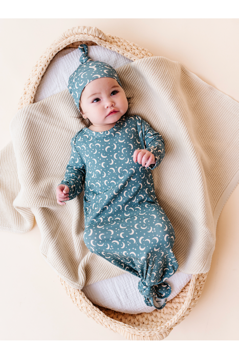 A baby wearing a dusty blue long-sleeve footed sleeper with a white crescent moon pattern and a matching pointed bamboo sleep hat, lying on a cream knit blanket inside a woven basket. Bird and Bean Baby 