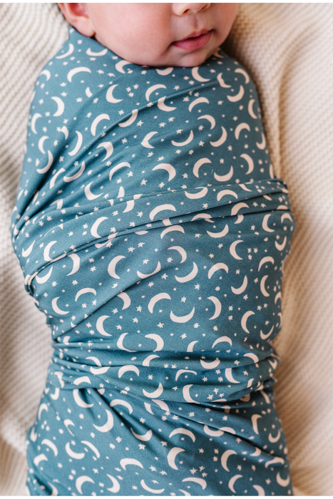 A close-up of a baby wrapped in a dusty blue bamboo swaddle blanket with a white crescent moon and star pattern, lying on a textured cream blanket. Bird and Bean Baby 