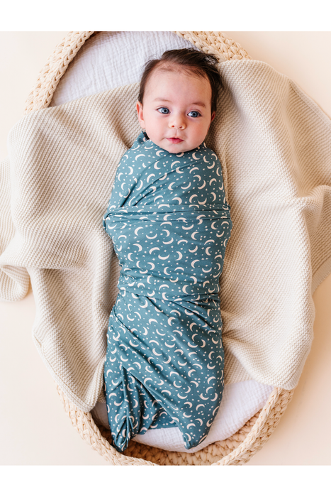 A baby swaddled in a dusty blue bamboo swaddle blanket with a white crescent moon pattern, lying inside a woven basket on a cream-colored blanket. Bird and Bean Baby 