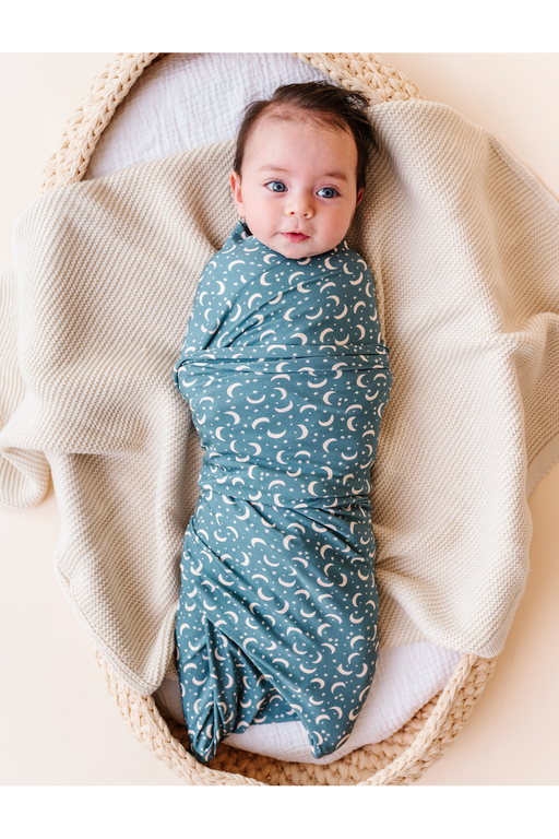 A baby swaddled in a dusty blue bamboo swaddle blanket with a white crescent moon pattern, lying inside a woven basket on a cream-colored blanket. Bird and Bean Baby 