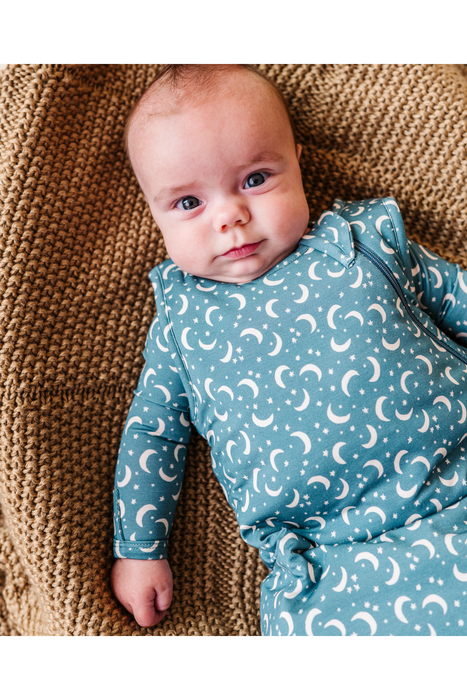 A baby lying on a brown knit blanket, wearing a long-sleeve footless bamboo TOG 1.0 sleep sack in dusty blue with a white crescent moon and star pattern. Bird and Bean Baby 