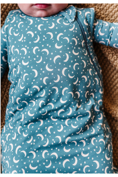 A close-up of a baby wearing a long-sleeve bamboo sleep sack in dusty blue with a white crescent moon and star pattern, lying on a textured brown blanket. Bird and Bean Baby Gifts 