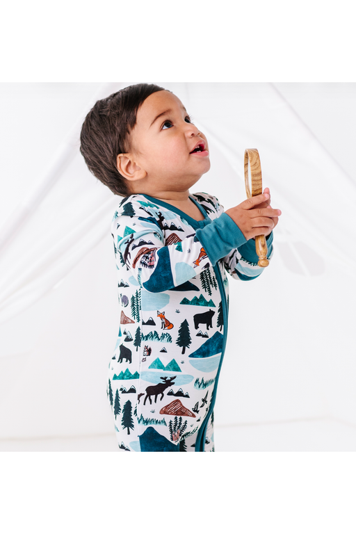 A toddler wearing a long-sleeve footless pajama zip set with a forest and mountain print, holding a wooden toy against a white studio background. Into the Wild Print by Bird and Bean