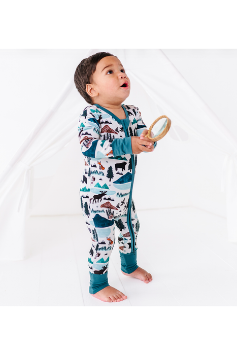 A toddler standing barefoot in a white tent-like setting, wearing a one-piece footed bamboo fabric pajama with a colorful animal and mountain print, and holding a small magnifying glass. Bird and Bean Kids, Into the Wild Print