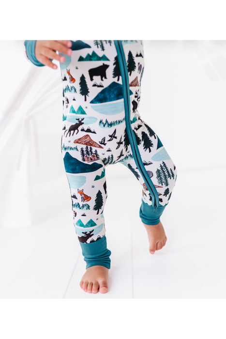 A close-up of a child wearing patterned winter pajamas with a front zipper. The pajamas have a white base with a colorful print of mountains, trees, and animals in teal, black, brown, and orange, with solid teal cuffs. Bird and Bean, into the Wild print 