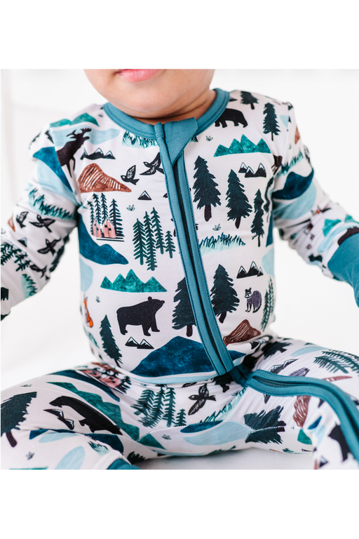 A close-up of a baby wearing a long-sleeve footless zip-up sleeper with a colorful forest and mountain print on a white background. The print features pine trees, bears, mountains, and cabins in shades of teal, black, brown, and blue. Into the Wild Print by Bird and Bean Kids