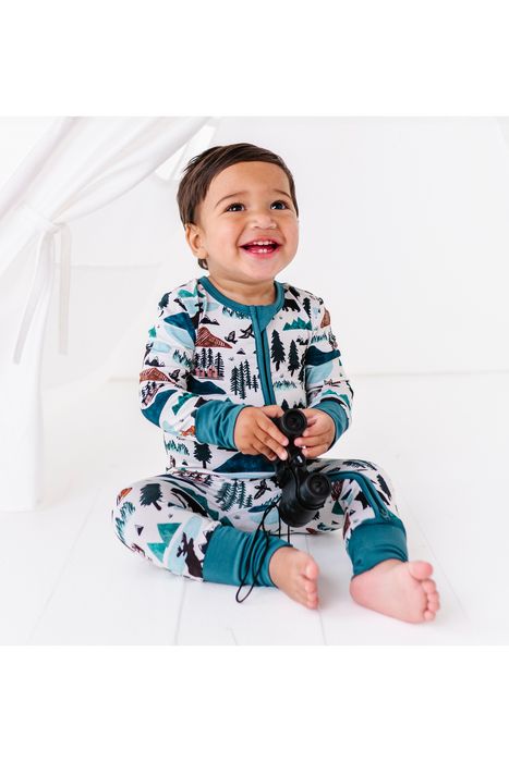 A smiling toddler sitting on a white floor wearing a long-sleeve zip-up pajama set with a forest and mountain print in teal, white, and brown. The child is holding a black toy binoculars. Into the Wild Print by Bird and Bean 