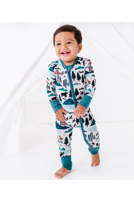 A smiling toddler stands barefoot in a white studio setting, wearing a one-piece zip-up pajama with a scenic outdoor print in teal, black, brown, and white. Into the Wild Bird and Bean Print 
