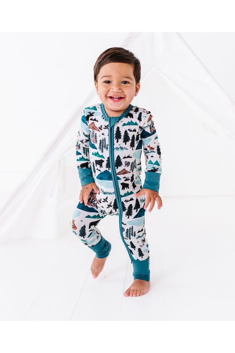 A smiling toddler wearing a full-zip one-piece pajama with a colorful mountain and forest pattern, standing barefoot against a white studio backdrop. Bird and Bean Kids 