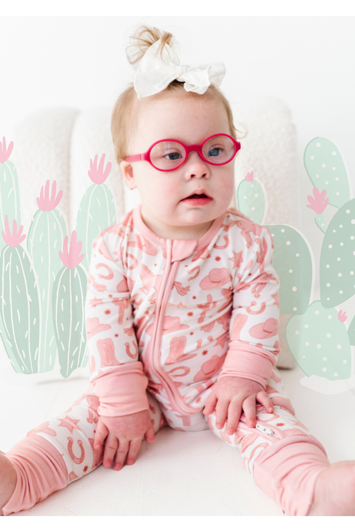 baby model wearing pink glasses wearing Bamboo Dixie Cowboy Western Pink Zip Romper sitting in studio with cactus designs layered behind