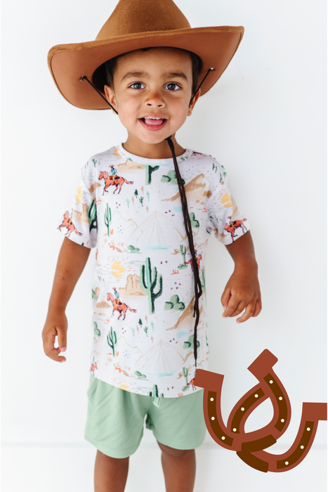 child model wearing billy western short and shirt set with cowboy hat on and graphic of two horseshoes overlaid on image